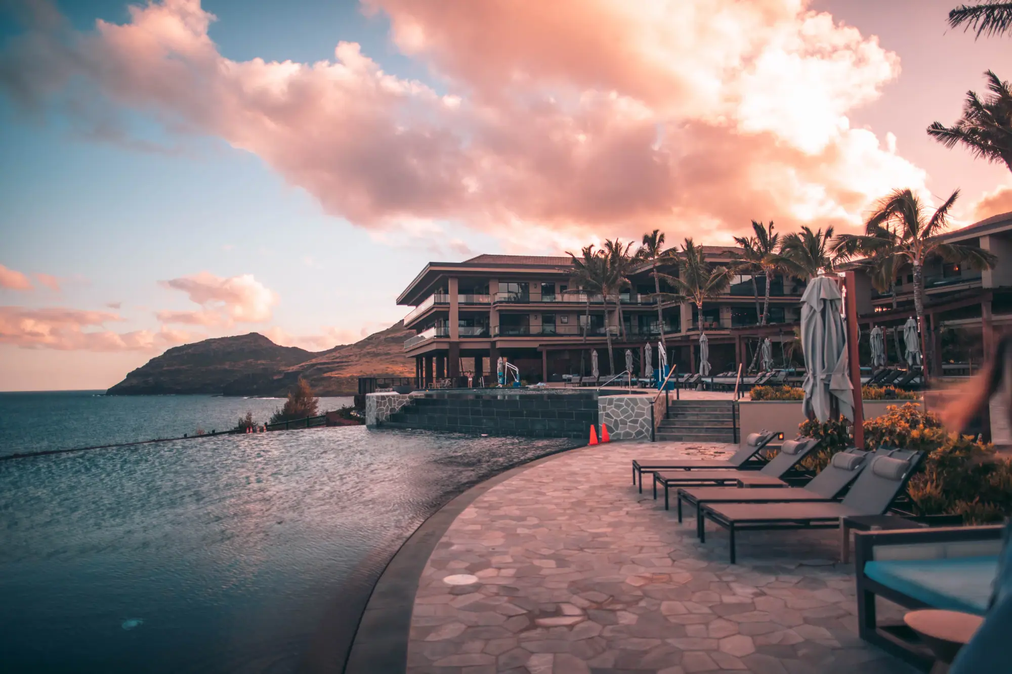 A resort infinity pool at the water's edge under a dramatic evening sky, loungers along the deck